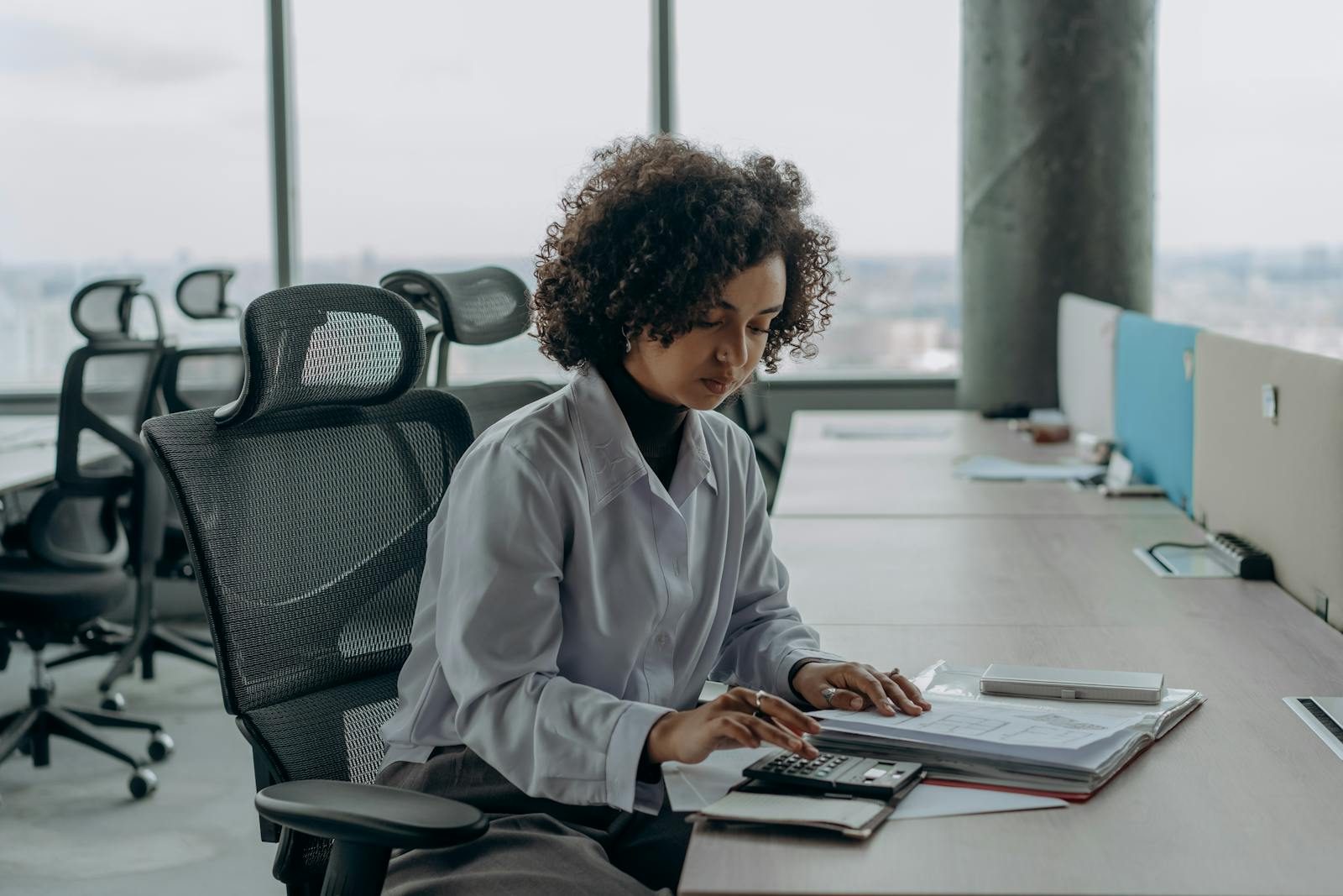 A professional woman with curly hair calculating documents in a modern office setting.